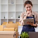 A woman in an apron stands among stacked shipping boxes, clipboard in hand, appearing to check inventory for her new Florida business.